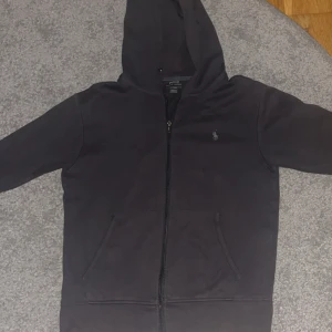 Mörkgrå hoodie från Polo Ralph Lauren - Mörkgrå hoodie från Polo Ralph Lauren med dragkedja framtill och klassisk huva. Tröjan har två fickor på magen och den ikoniska lilla loggan broderad på bröstet. Perfekt för en chill och stilren look.( Inga snören)