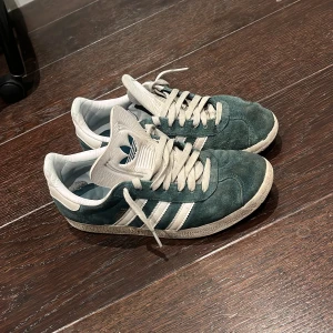 Adidas Gazelle gröna sneakers mocka - Säljer ett par klassiska Adidas Gazelle sneakers i mörkgrön mocka med vita detaljer och snörning. Skorna har den ikoniska tre-randiga designen på sidorna och vit logga på tungan och hälen. Perfekta för dig som gillar retrostil och streetwear.