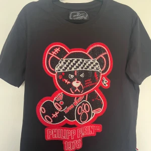 Svart Philipp Plein t-shirt med björn - Svart t-shirt från Philipp Plein med stor, detaljerad björn i rött, vitt och silver framtill. Björnen har coola detaljer och texten 'PHILIPP PLEIN 1978' under sig. Klassisk passform och rund hals. Perfekt statement-plagg för dig som gillar streetstyle.