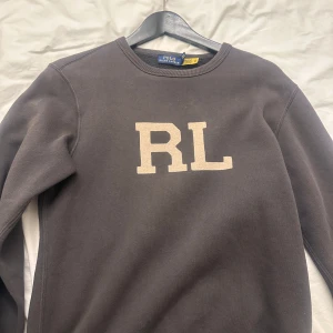 Grå sweatshirt från Polo Ralph Lauren - Mörkgrå sweatshirt från Polo Ralph Lauren med stora vita RL-bokstäver på bröstet. Klassisk rund halsringning och långa ärmar. Perfekt för en avslappnad och stilren look.