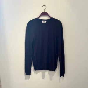 Svart stickad tröja från NN07 i ull - En stilren svart stickad tröja från NN07 i storlek M som är i 100% fin ull. Tröjan har rund halsringning, långa ärmar och ribbade muddar vid ärmslut och nederkant. Perfekt för lager-på-lager och enkel att matcha med allt. Hör av er vid frågor