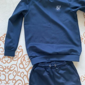 Mörkblå tröja från SikSilk set - Mörkblå långärmad tröja från SikSilk med rund hals och vit broderad logga på bröstet. Tröjan är i mjukt material och har ribbade muddar vid ärmslut och nederkant. Perfekt för en chill och sportig stil. Byxorna har samma stilrena design med logga framtill, resår i midja och benslut.