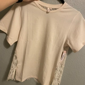 Beige topp med spetsdetaljer XS - HELT NY: Superfin beige topp från Nelly i storlek XS. Framsidan är i mjuk bomull och baksidan har genomskinlig spets med blommigt mönster. Korta ärmar och rund halsringning. Perfekt för dig som gillar romantiska detaljer! 