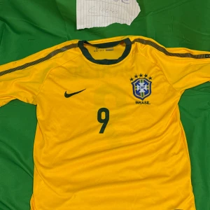 Brasilien Ronaldo 9 Nike fotbollströja - Gul Brasilien fotbollströja från Nike med gröna detaljer, nummer 9 och Ronaldo på ryggen. Klassisk landslagströja med CBF-emblem och Dri-Fit-material som andas. Perfekt för dig som älskar fotboll och vill hylla en ikon.Liknande modell som originalet!