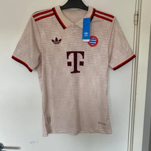 Bayern München Adidas fotbollströja - Snygg Bayern München fotbollströja från Adidas med klassiskt klubbmärke och T-Mobile-logga. Tröjan är beige med röda detaljer och diskret mönster, har krage och röda ränder på axlarna. Tillverkad i lätt och ventilerande material.