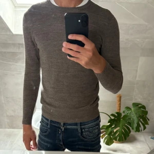 Brun merinoullströja från A Day's March - Säljer en stilren brun tröja i merinoull från A Day's March. Tröjan har rund halsringning, ribbade muddar och är långärmad. Perfekt för lager-på-lager och funkar året runt. Enkel och snygg design som passar till jeans eller chinos.