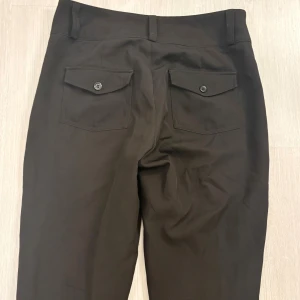 Svarta kostymbyxor med fickor - Snygga svarta kostymbyxor från Gina tricot med raka ben och två bakfickor med lock och knapp. Byxorna har bälteshällor och är tillverkade i ett mjukt tyg som ger en dressad look. Väldigt slitna längst ner (kan skicka bilder) pga trampat på de men övrigt i bra skick. 