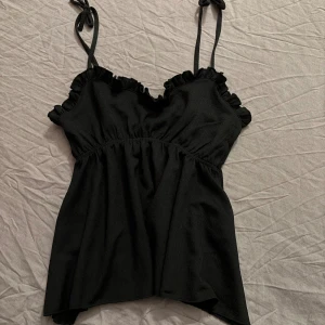 Svart linnetopp med volang och knytband - Säljer en svart linnetopp med smala axelband som knyts upptill och volangdetaljer längs urringningen. Toppen har en lätt utställd passform och resår under bysten för en söt och avslappnad look.