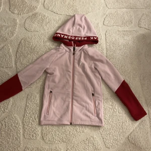 Rosa hoodiejacka Peak Performance - Ljusrosa hoodiejacka från Peak Performance med röda ärmslut och röd insida på huvan där loggan står tryckt. Jackan har dragkedja framtill och två fickor med dragkedja. Perfekt för en sportig och chill stil.