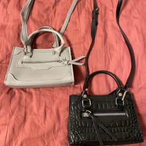 Två små handväskor i svart och grått - Säljer två stilrena små handväskor med axelrem, en i ljusgrått och en i svart med croco-mönster. Båda har dragkedja framtill och snygga detaljer. Perfekta och tuffa. Aldrig använda