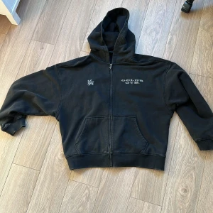 Svart Gold's Gym hoodie från YoungLA - Svart hoodie från YoungLA med stor silvrig Gold's Gym-logga på ryggen och mindre tryck fram. Dragkedja hela vägen, huva och känguruficka. Snygga detaljer på ärmen och bröstet. Perfekt för gymmet eller chill.