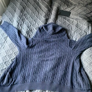 Mörkblå hoodie från Polo Ralph Lauren - Mörkblå kabelstickad hoodie från Polo Ralph Lauren med dragkedja, huva och fickor framtill. Klassisk röd logga broderad på bröstet. Tröjan har långa ärmar och ribbade muddar för en snygg passform.