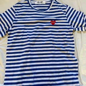 Randig t-shirt från Comme des Garçons - Säljer en blåvit randig t-shirt från Comme des Garçons PLAY med det ikoniska röda hjärtat broderat på bröstet. T-shirten har korta ärmar och är gjord i mjuk bomull. Perfekt för dig som gillar streetstyle och vill sticka ut lite extra.