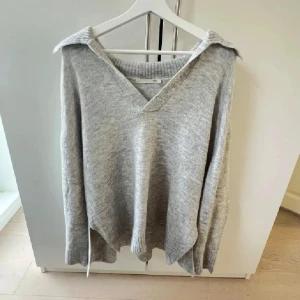 Grå stickad v-ringad tröja - Mysig grå stickad tröja med v-ringning och bred krage. Tröjan har en avslappnad passform och långa ärmar, perfekt för lager på lager.