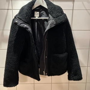 Svart pile/Teddy Jacka från H&M - Mysig svart teddy/pile jacka från H&M i storlek XS. Jackan har hög krage, två stora fickor framtill och stängs med dragkedja och tryckknappar. Perfekt för kalla dagar och ger en cool, avslappnad vibe.