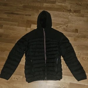 Svart pufferjacka med huva - Svart pufferjacka  med huva och broderad logga på ryggen. Jackan har quiltad insida, dragkedja framtill och två sidofickor med dragkedja. Elastisk nederkant med logo-tryck. Perfekt för kalla dagar och snygg streetstil. Den va för liten för mig så kan även tänka mig byta mot en annan jacka i XL. Denna är i M/L. Nfc tagg finns. 