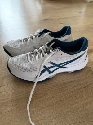 Asics Gel-Rocket 12 White/Mako Blue - Säljer ett par Asics Gel-Rocket 12 sneakers i vitt och mörkblått. Skorna har meshpaneler för ventilation, klassisk snörning och en platt sula. Snygg sportig design med Asics-loggan på sidan och bekväm passform, perfekta för träning eller sport. Helt oanvända.