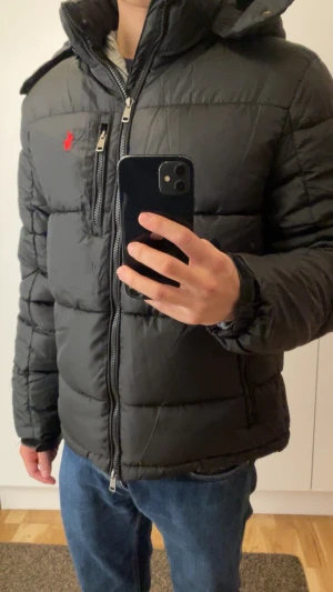 Svart pufferjacka från Polo Ralph Lauren - Svart pufferjacka från Polo Ralph Lauren med röd logga på bröstet. Jackan har huva, tvåvägsdragkedja med RL-detalj, quiltad design och flera fickor med dragkedja. Perfekt för kalla dagar och har en snygg, sportig look.