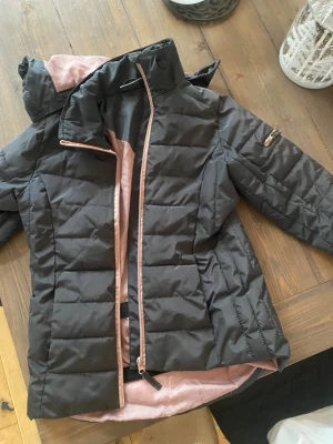 Svart quiltad pufferjacka med rosa detaljer - Snygg svart pufferjacka med quiltad design och ljusrosa foder samt dragkedja. Jackan har hög krage, huva och långa ärmar. Perfekt för kyliga dagar och ger en cool look med sina kontrastdetaljer.