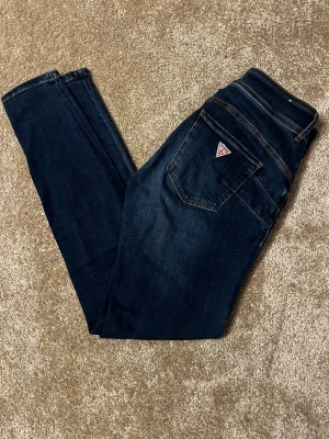 Guess Skinny High Shape Up jeans - Mörkblå skinny jeans från Guess med hög midja. Modellen heter Skinny High Shape Up med diskret logga på bakfickan. Jeansen är tillverkade i stretchigt denim för en tight och bekväm passform. Passar storlek 36-42.