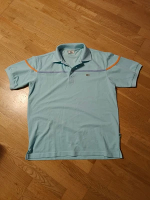 Ljusblå piké från Lacoste - Snygg ljusblå pikétröja från Lacoste med klassisk krage och korta ärmar. Tröjan har orange och lila detaljer på axlarna och en liten krokodillogga på bröstet. Tillverkad i mjuk bomull som är skön mot huden. Perfekt för dig som gillar sportig och fräsch stil.