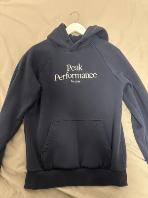 Marinblåa peak performance hoodie  - En marinblåa peak performance hoodie. Hoodien håller fortfarande sin blåa färg och är i gott skick, ser använd ut men inte förstörd eller trasig. Koftan är köpt på Johnells och är i storlek S. Snören finns kvar men jag har plockat ut dom, om de önskans kan jag skicka med dom. 