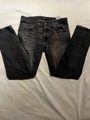Svart/Gråa tapered jeans från Jack & Jones - Snygga svarta jeans från Jack & Jones i modellen Tapered/Mike. Jeansen har en tvättad look med klassiska fem fickor, knappgylf och smalare ben. Perfekt för dig som gillar en modern och avslappnad stil. Storlek 32/32.