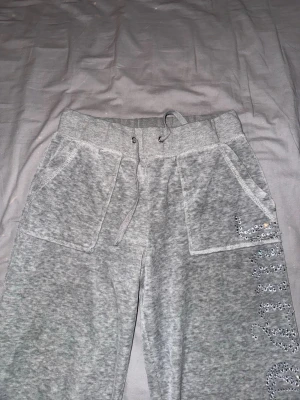 Grå mjukis set från Juicy Couture XS - Säljer ett grått set från Juicy Couture med glittriga paljettdetaljer längs ena byxsidan och backsidan på tröjan. har breda fickor fram och mindre bak, snörning i midjan och mjukt, bekvämt material. Perfekt för dig som gillar chill och glam i ett.