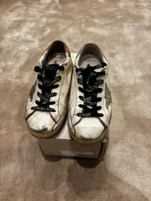 Golden Goose vita sneakers med glitter - Golden Goose sneakers i vitt skinn med svart snörning och silverfärgad stjärndetalj på sidan. Hälpartiet har glittrig guldton och platt sula. Snyggt slitna med cool vintage vibe, perfekt för dig som gillar statement-skor med unik look.