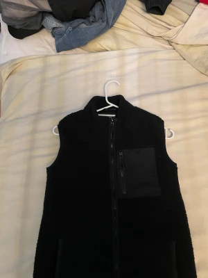 Svart fleeceväst med dragkedja - Svart fleeceväst med hög krage och hel dragkedja framtill. Västen har en praktisk bröstficka med dragkedja i ett slätt material och två sidofickor. Perfekt för lager-på-lager och chill stil. Enkel, stilren och riktigt skön att bära.