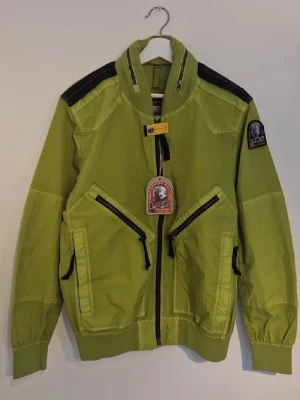 Grön vindjacka från Parajumpers - Snygg grön vindjacka från Parajumpers med svarta detaljer på axlarna och dragkedjor på fickorna. Jackan har hög krage  och klassiska parajumper-emblem på ärmen. Perfekt för dig som vill sticka ut med en cool och funktionell look.