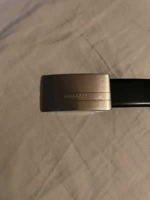 Svart Hugo Boss skärp i skinn - Stilrent svart skärp från Hugo Boss med en snygg, matt silverfärgad metallspänne där loggan är ingraverad. Skärpet är tillverkat i äkta skinn och har en klassisk, rak form som passar till jeans eller kostymbyxor. Perfekt accessoar för en clean look. 105cm långt 85/32