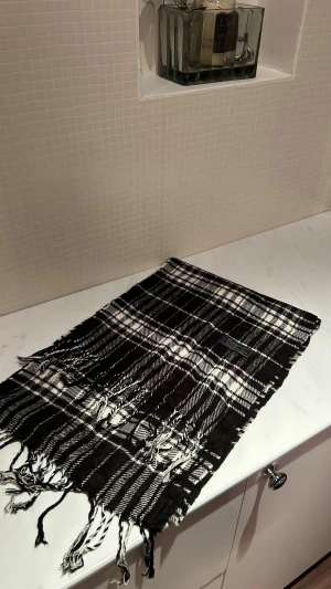 Svartvit rutig ullväst från Burberrys - Snygg svartvit rutig väst från Burberrys London, tillverkad i 100% lammull. Klassisk design med fransar längs nederkanten och ett tydligt Burberrys-märke. Perfekt för dig som gillar tidlös stil och vill ha något som sticker ut.