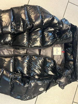 Svart Moncler pufferjacka - Säljer en svart, glansig pufferjacka från Moncler med klassisk logga på fickan. Jackan har quiltad design, dragkedja framtill och stora fickor med lock. Perfekt för dig som vill ha en snygg och varm jacka med street vibe.
