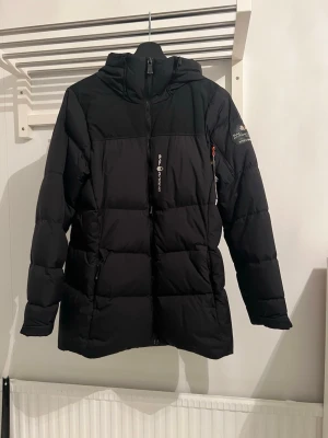 Svart Sail Racing dunjacka med huva - Säljer en svart dunjacka från Sail Racing med huva och snygga detaljer som patch på ärmen och logga på bröstet. Jackan har dragkedja framtill och två sidofickor med dragkedja. Perfekt för kalla dagar och har en stilren, sportig look.