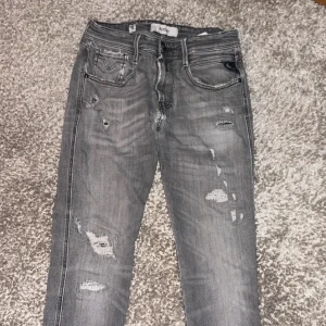 Grå slitna jeans från replay  - Snygga grå jeans från replay med slitningar och trasiga detaljer på både fram- och baksida. Klassisk femficksmodell med svarta broderier på bakfickorna och läderpatch i midjan. Perfekt för dig som gillar streetwear och en rå look. Modell anbass, OBS 2 hål i bak