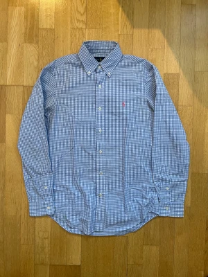 Blårutig skjorta från Ralph Lauren XS - Snygg blårutig skjorta från Ralph Lauren i storlek XS. Skjortan har klassisk krage, långa ärmar och knappar framtill. Tillverkad i 100% bomull och har en liten rosa broderad logga på bröstet. Perfekt för dig som gillar stilrena och tidlösa plagg.