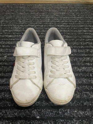 Sneakers - Vita sneakers från H&M med röda och blå ränder på sidan och röd häl med blå ögla. Skorna är unisex, i storlek 33 och inte jättemycket använda.