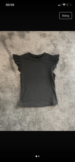 Svart topp med volangärmar från SHEIN - Svart topp från SHEIN i storlek S med rund hals och korta ärmar dekorerade med snygga volanger. Modellen är croppad och har en stilren, enkel design som passar till många olika looks. Materialet känns mjukt och lätt, perfekt för dig som gillar trendiga detaljer.