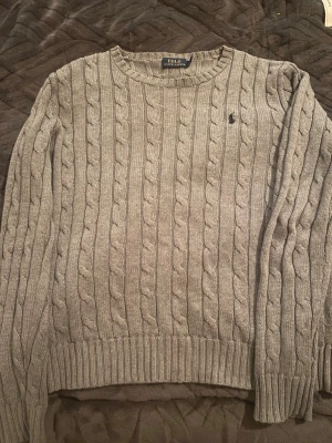 Grå kabelstickad tröja Polo Ralph Lauren - Snygg grå kabelstickad tröja från Polo Ralph Lauren med rund hals och diskret logga broderad på bröstet. Tröjan har långa ärmar och ribbade muddar vid ärmslut och nederkant. Perfekt för dig som gillar klassisk och stilren look. Höjden på tröjan är 50 cm och bredden är 37 cm 😌