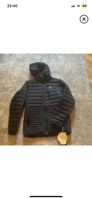 Svart dunjacka från Haglöfs med huva - Svart dunjacka från Haglöfs med huva och snygga horisontella quiltade paneler. Jackan har dragkedja framtill, bröstficka med logga och två sidofickor. Perfekt för kalla dagar och har en clean, sportig look som funkar till streetwear. Passar även S se sista bilden. Perfekt till detta kalla väder