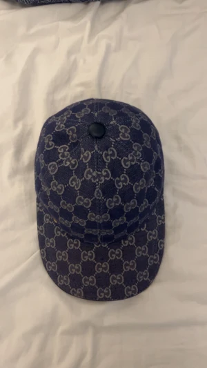 Gucci blå keps med GG-mönster - Snygg blå keps från Gucci i denimliknande tyg med klassiskt GG-mönster över hela. Justerbar rem bak i mörkblått läder med guldfärgad spänne. Svart knapp på toppen och ljus insida med etikett. Perfekt för dig som gillar lyxiga streetwear-accessoarer.
