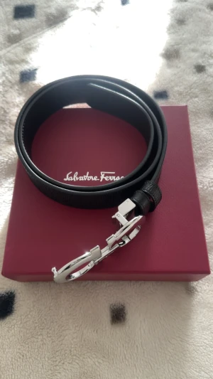 Svart bälte från Salvatore Ferragamo - Exklusivt svart bälte från Salvatore Ferragamo med en stilren, blank silverfärgad spänne i deras ikoniska Gancini-design. Bältet är tillverkat i äkta skinn med en lätt texturerad yta och har en klassisk, rak form. Perfekt för dig som gillar lyxiga accessoarer med italiensk touch. Priset kan diskuteras 