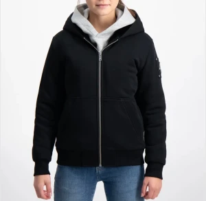 Svart bomberjacka från Moose Knuckles - Svart bomberjacka med huva från Moose Knuckles, har en snygg metall-logga på ärmen och praktiska fickor framtill. Jackan är tillverkad i mjuk bomull och polyester, med dragkedja och ribbade muddar. Perfekt för dig som gillar streetstyle och vill ha en clean look. Den är i str ” L ” barn storlek och passar de som är 157 - 165 cm