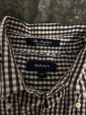 Rutig skjorta från GANT, regular fit - Snygg rutig skjorta från GANT i regular fit. Skjortan är vit och brun/mörklila med klassiskt rutmönster, har knappar framtill och bröstficka med broderad GANT-logga. Tillverkad i mjuk bomull och har långa ärmar. Perfekt för en clean och stilren look.