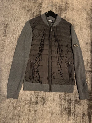 Barbour cardigan - Grå cardigan från Barbour med quiltad front och gråa ärmar. Jackan har ribbade muddar, dragkedja framtill och två fickor med zip.