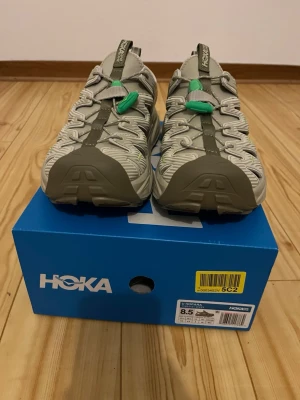 Hoka Hopara sandaler beige/grön - Säljer ett par Hoka Hopara sandaler i beige med gröna detaljer och robust platt sula. Skorna har öppna partier på sidorna, snörning med gröna snörlås och dragflik bak. Tillverkade i syntetmaterial, perfekta för sommarens äventyr.
