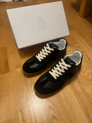 Maison Margiela sneakers i svart - Sneakers i svart med vita snören och gummisula. Klassisk low-top siluett med ovandel i skinn och mocka. Snygg minimalistisk design med diskreta detaljer och vit insida. Perfekta för dig som gillar stilrena och exklusiva sneakers.