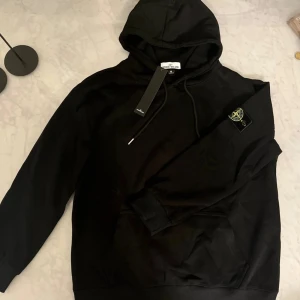 Svart hoodie från Stone Island M - Svart hoodie från i storlek M med klassisk logotyp-patch på ärmen. Tröjan har huva med snörning, ribbade muddar och en stor magficka. Materialet är mjuk bomull och passformen är normal. Perfekt för dig som gillar streetwear och snygga detaljer.