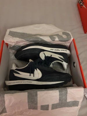 Nike LDWaffle Fragment x Sacai blå/vit - Nike LDWaffle Fragment x Sacai sneakers i mörkblå och vit färg med dubbla swoosh-loggor och textdetaljer på mellansulan. Skorna har en platt, räfflad sula och är tillverkade i mocka och syntetmaterial. Snygg retrostil med snörning och rund tå.